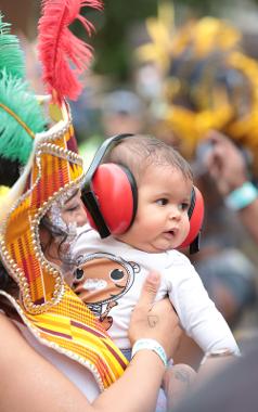 Woman in 2017 parade, dressed in carnival costume holding baby with ear defenders.