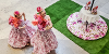 Two performers in pink floral dresses dance around table of tea and cakes