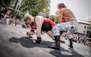 Two wrestlers in a ring with a referee in a wrestling ring on the Flag Market with crowds watching