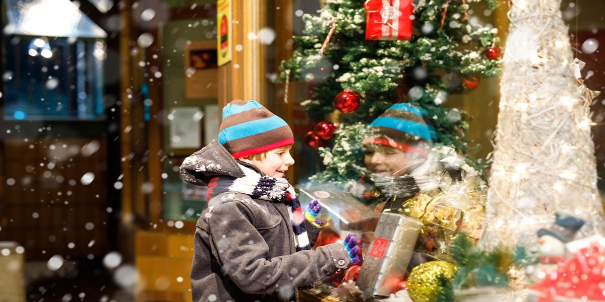 Funny happy child in fashion winter clothes making window shopping decorated with gifts, Xmas tree