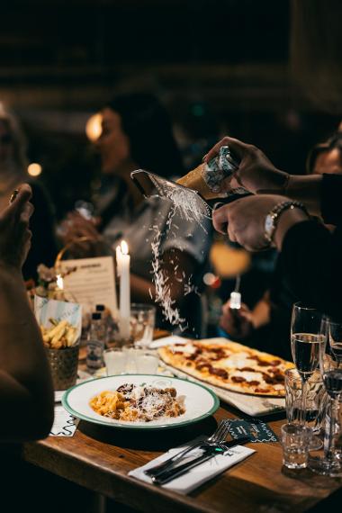 A person off camera grating cheese over a plate of pasta on a guests table