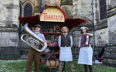 Three people holding instruments stood in front of a cart that reads The Guild of Toasters