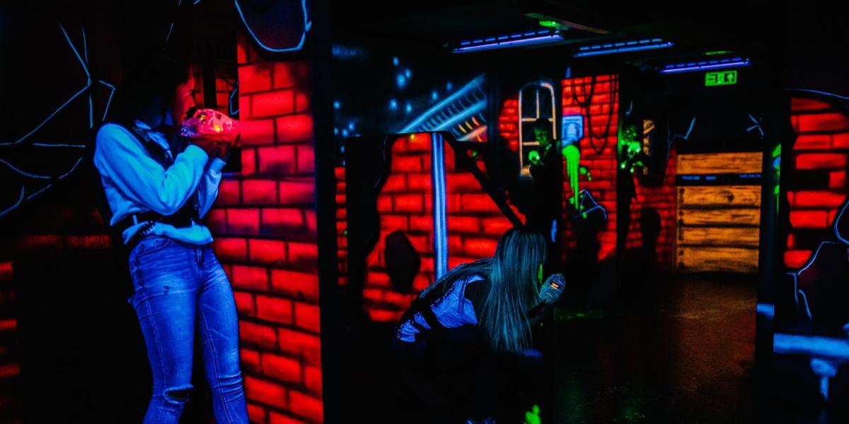 Two girls hiding behind End of Days laser tag arena walls, lit by UV light.