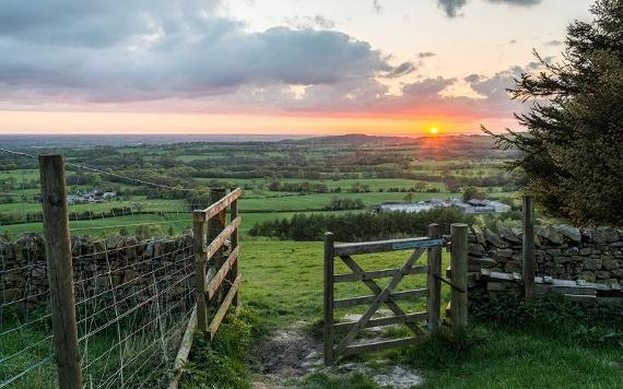 Gate open leading onto field with view over more fields and sunset.