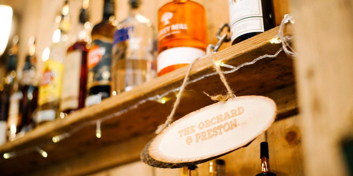 Main image: Wooden sign for The Orchard hanging by spirits behind bar. Wooden sign for The Orchard hanging by spirits behind bar.