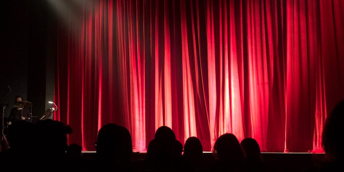 Main image Initiate Theatre Audience sat in theatre with red curtain closed on stage.