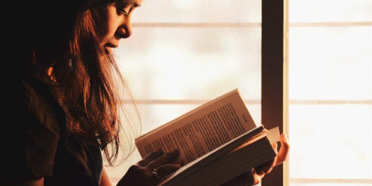 Woman reading book by window in low light.